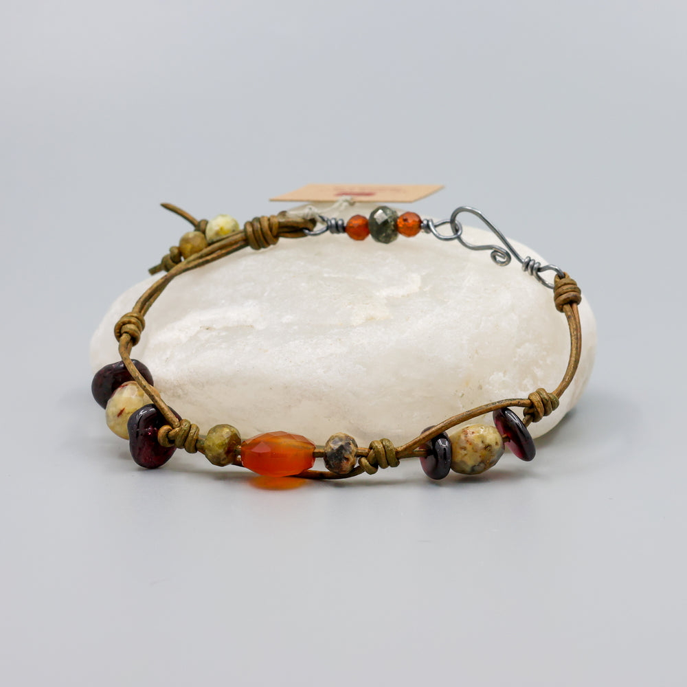 handmade leather and gemstone bracelt with garnet, carnelian, crazy lace agate, and hessonite with oxidized sterling silver clasp shown on a crystal rock