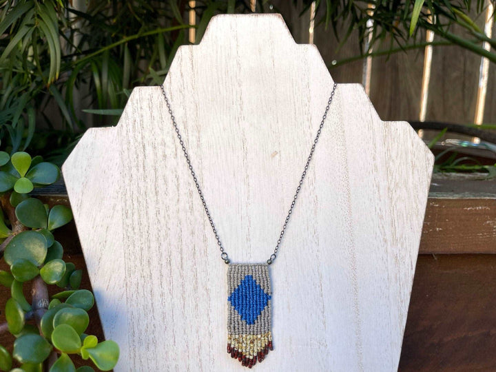 Handwoven four elements macrame necklace in blue and tan with tan and red seed bead fringe on a white background