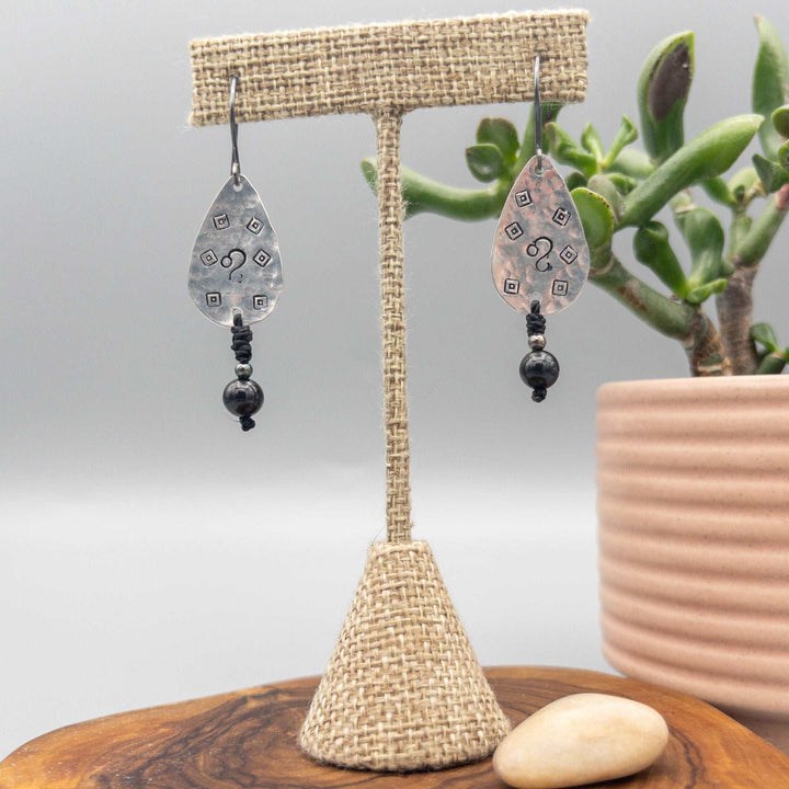 Hand-stamped Leo earrings with black spinel beads hanging from a linen dieplay