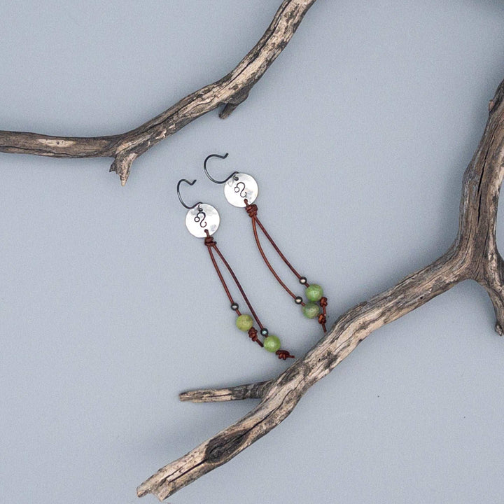 Sterling silver Leo stamped earrings with peridot beads on long leather cord flat on gray