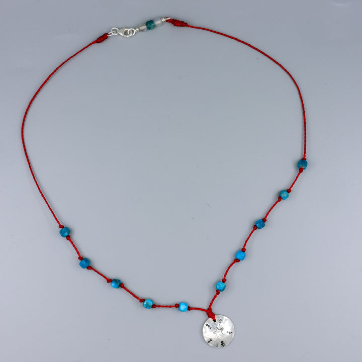 handmade silver stamped necklace with blue apatite beads on a red cord on a gray background